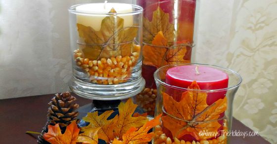 Three hurricane popcorn table decorations on a table with leaves and pinecones around them.