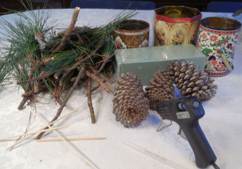 Supplies for DIY pine cone Christmas trees including fir branches, pine cones, decorative tins, floral foam and a glue gun.