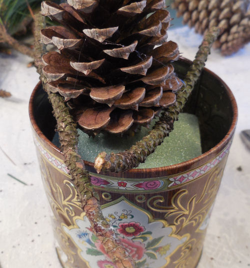 A pine cone is propped up on some floral foam by sticks to help the glue dry.