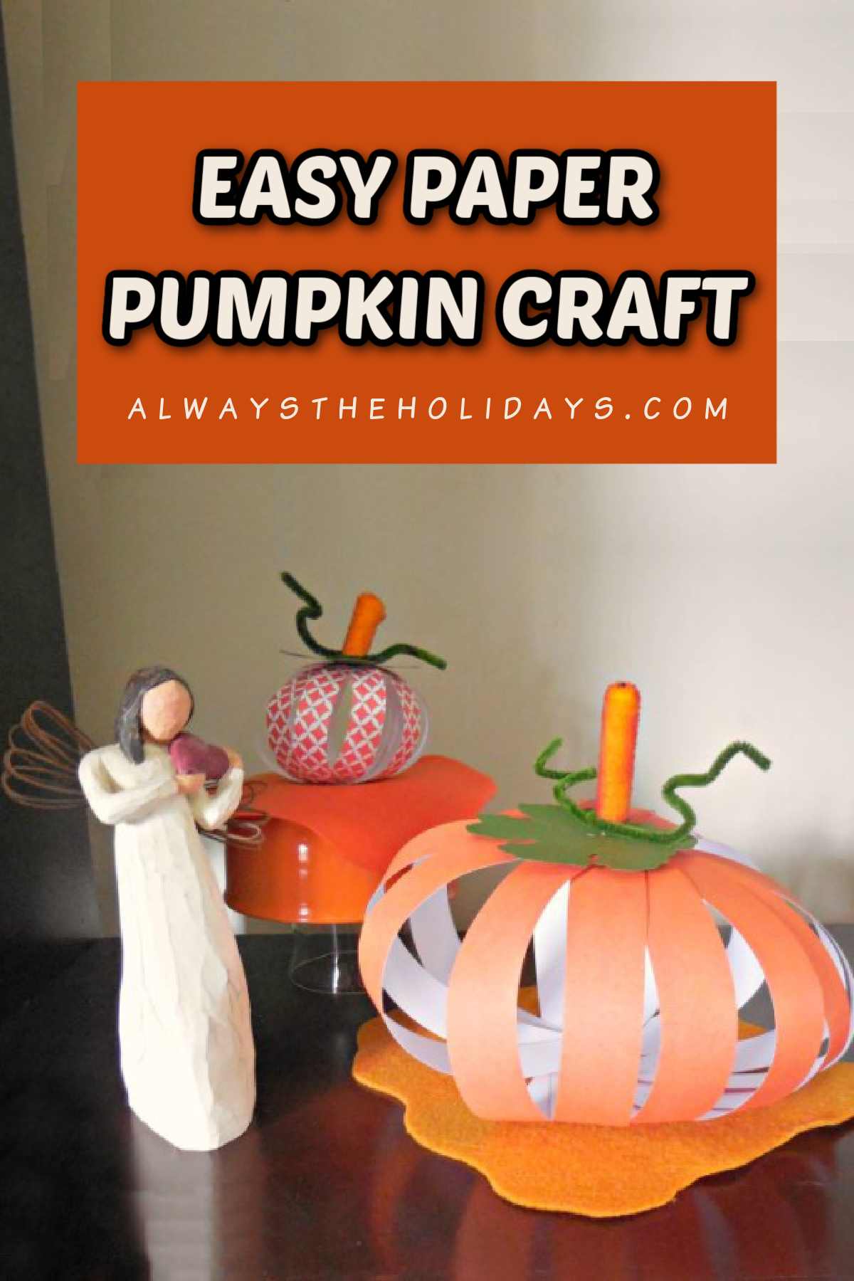 A simple paper pumpkin craft displayed on a mahogany table between two angel figurines with a text overlay that says DIY scrapbook paper pumpkins in white and orange lettering.