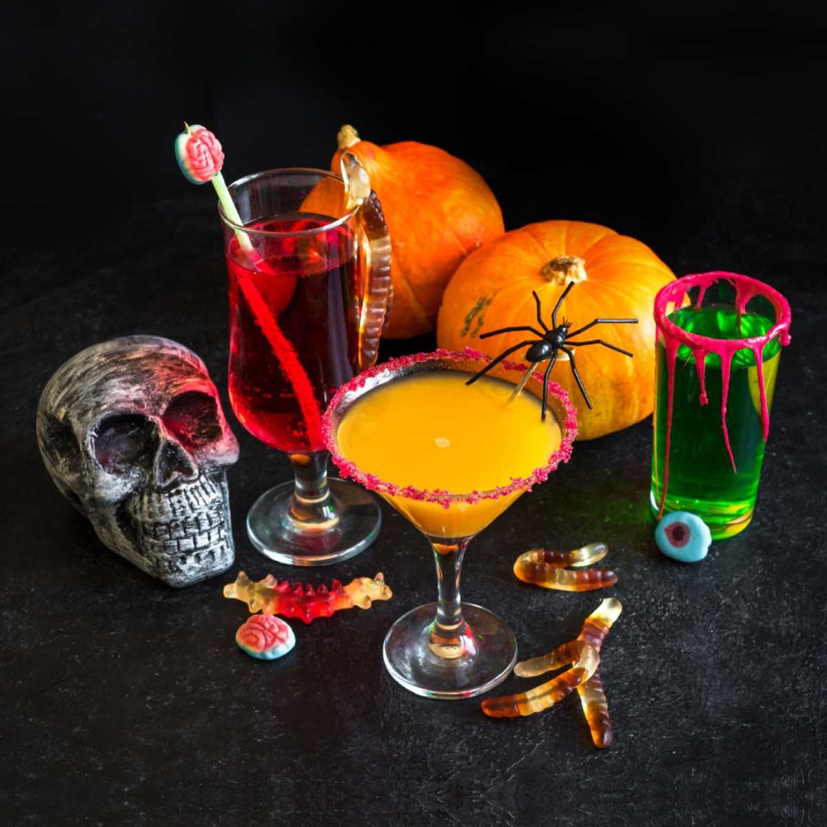 An assortment of Halloween drinks for adults, with various candy garnishes beside a silver skull and two small pumpkins.