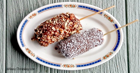 Two Paleo and gluten-free frozen banana pops on a white plate with a blue border. One of the banana pops is coated with pecans, and the other is coated with coconut.