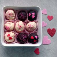 Nine black and white raspberry truffles in a porcelain dish next to paper hearts on a table.