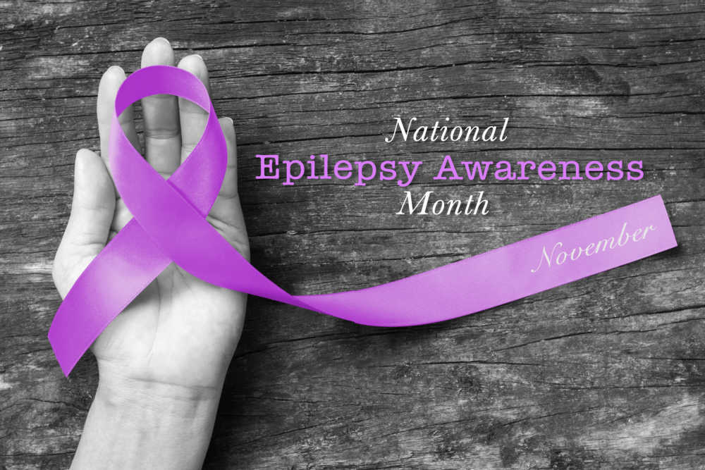 Hands holding a purple ribbon and words National Epilepsy Awareness Month - November.