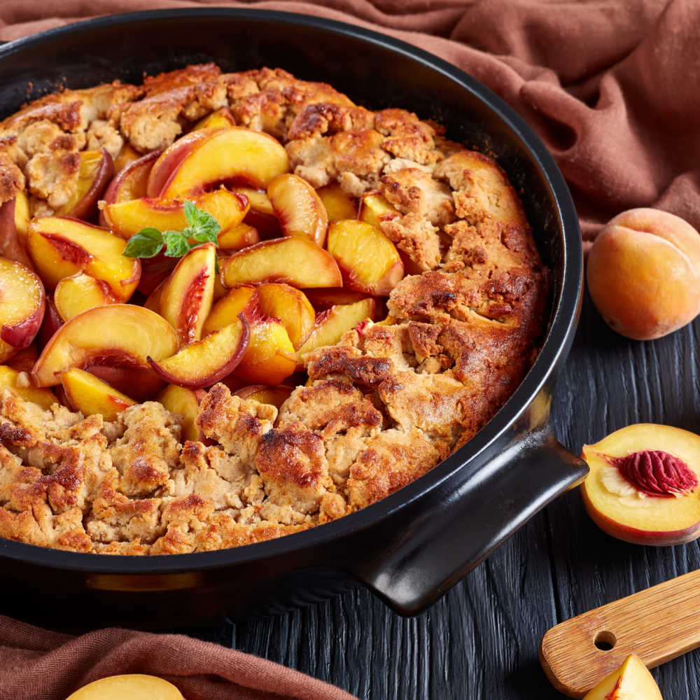 A photo of a peach cobbler recipe made in a cast iron pan, with a red cloth and peaches styled around it.
