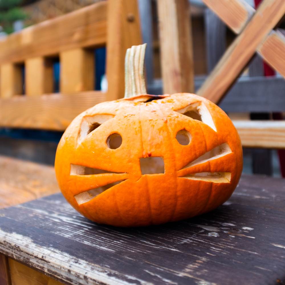 A geometric cat face pumpkin on a wooden table against a backdrop of more wooden furniture.