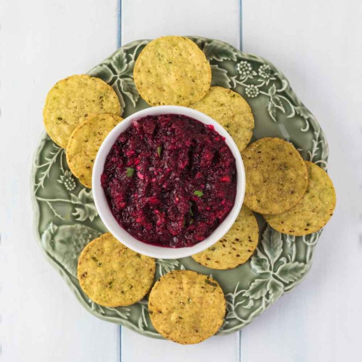 A white ramekin filled with an easy cranberry relish recipe sitting on a green plate surrounded by tortilla chips.