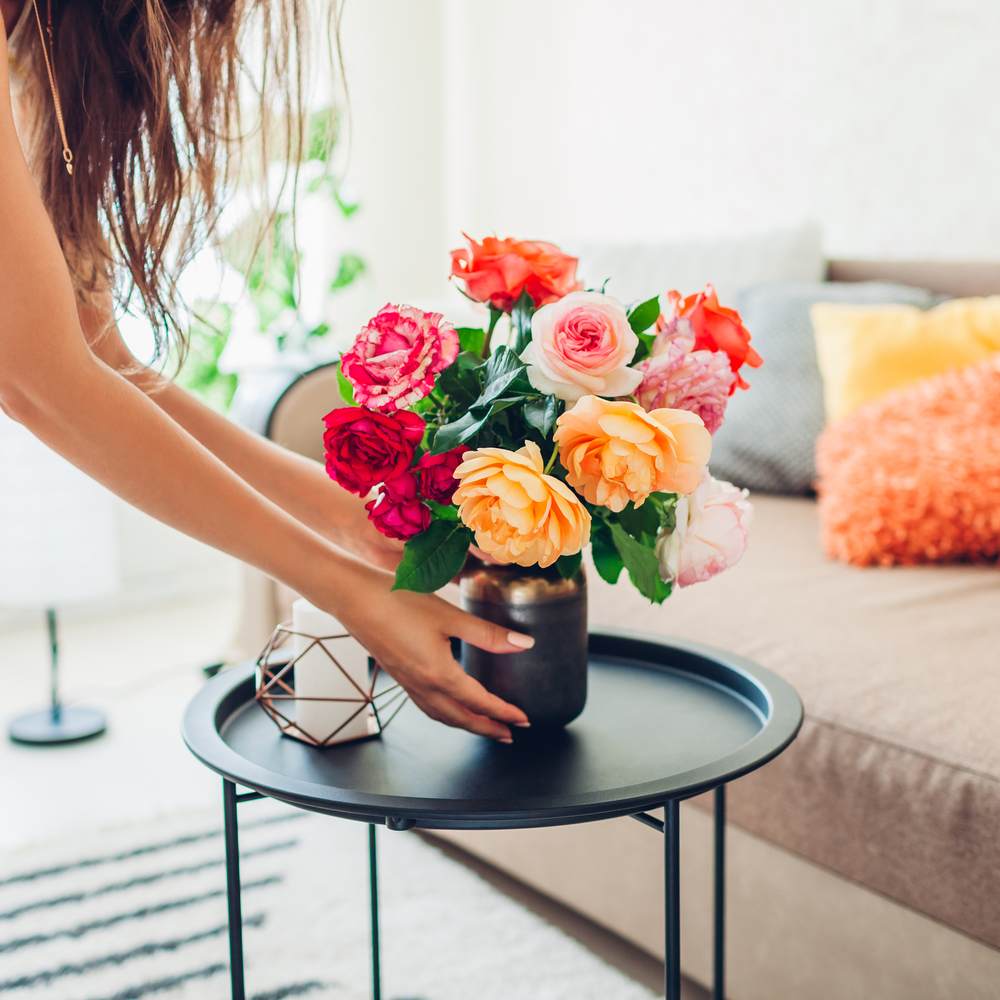 A woman putting a vase of roses on a side table in the living room, the vase has a rose of every color.