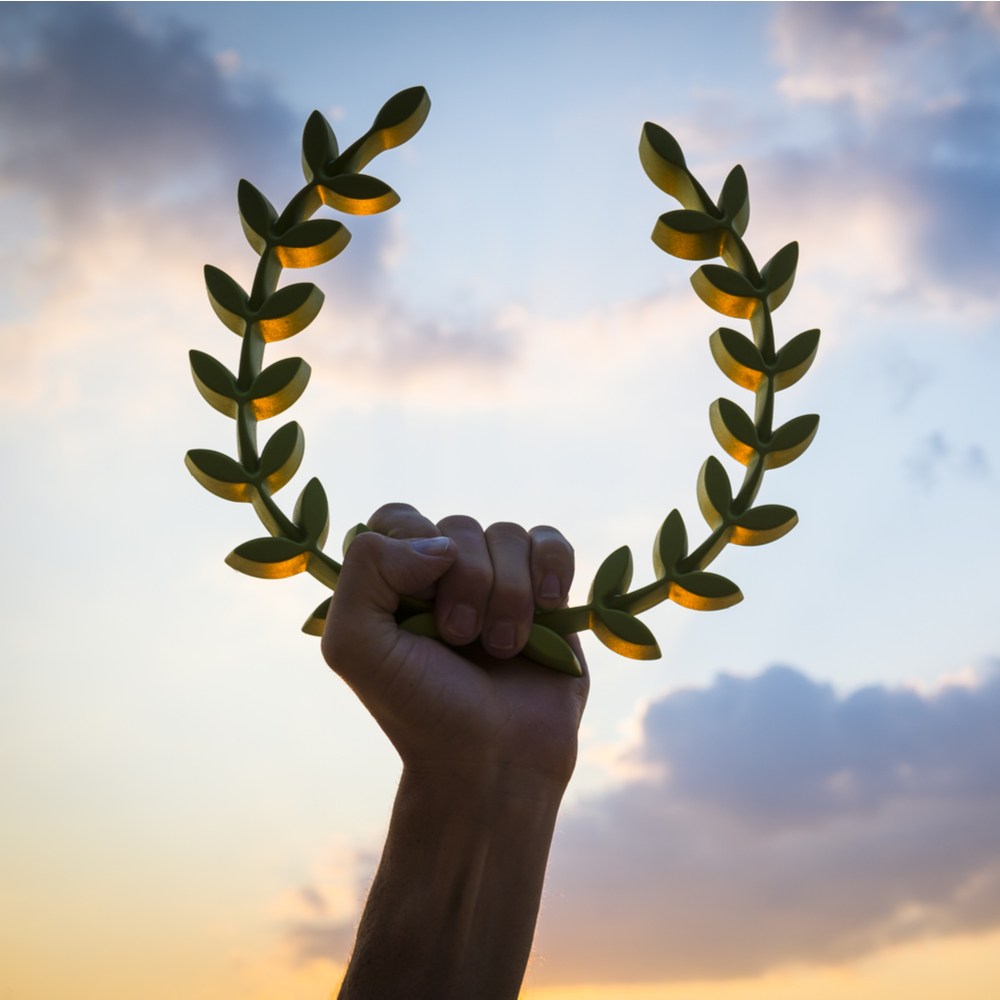 A hand holding a laurel wreath, one of the symbols of Apollo against a sky background that where the sun is setting and the sky is changing colors from blue to orange.