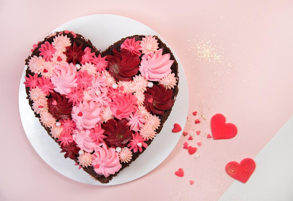 A chocolate cake make in the shape of a heart, one of the most popular Valentine's Day symbols with frosting in different shades of pink and red making icing flowers on a plate on a pink background with paper cut out hearts next to it.