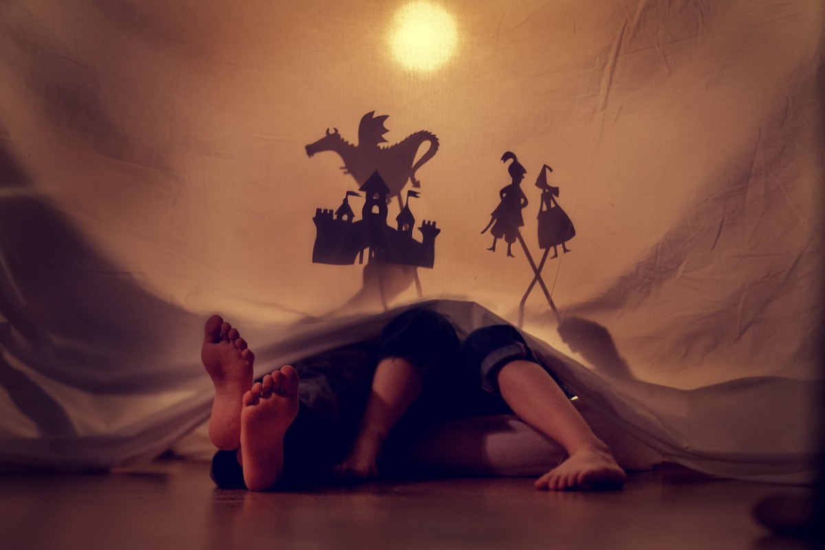 A parent and child behind a sheet with their feet sticking out of it, playing with shadow puppets, including a castle, a dragon, a prince and princes to retell a Greek mythology love story and fairy tale.
