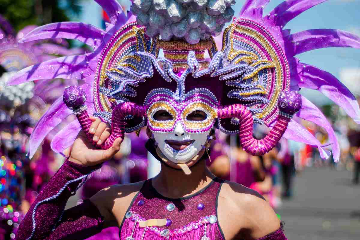 A krewe member marching in a Mardi Gras parade, wearing a full Mardi Gras mask that is purple and pink.