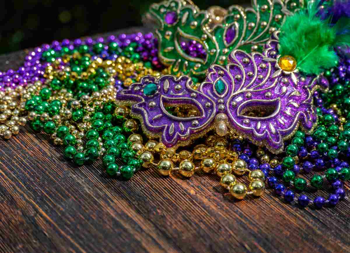 Mardi Gras beads and a Mardi Gras mask on a wooden table to honor the classic Mardi Gras traditions.