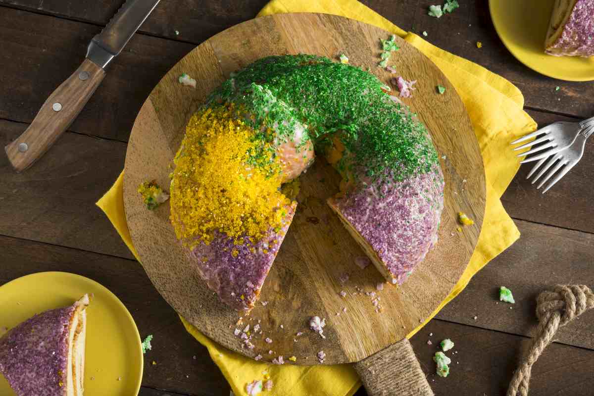 A king cake with purple, green and gold sprinkles laying on a circular cutting board with a slice missing and a yellow towel in the frame.