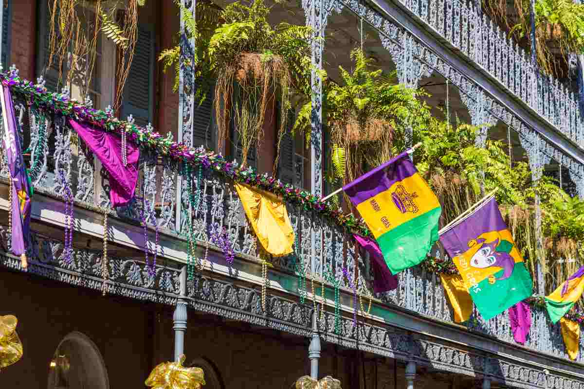 A balcony in the french quarter during the Mardi Gras parade with Mardi Gras flags waiving from the balcony, and Mardi Gras beads hanging from it.