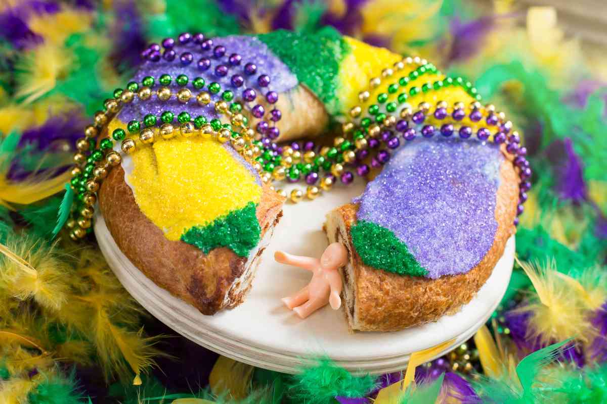 A king cake with purple, gold and green sprinkles, draped with Mardi Gras beads and a baby from the king cake sitting next to the cake where a slice is missing.