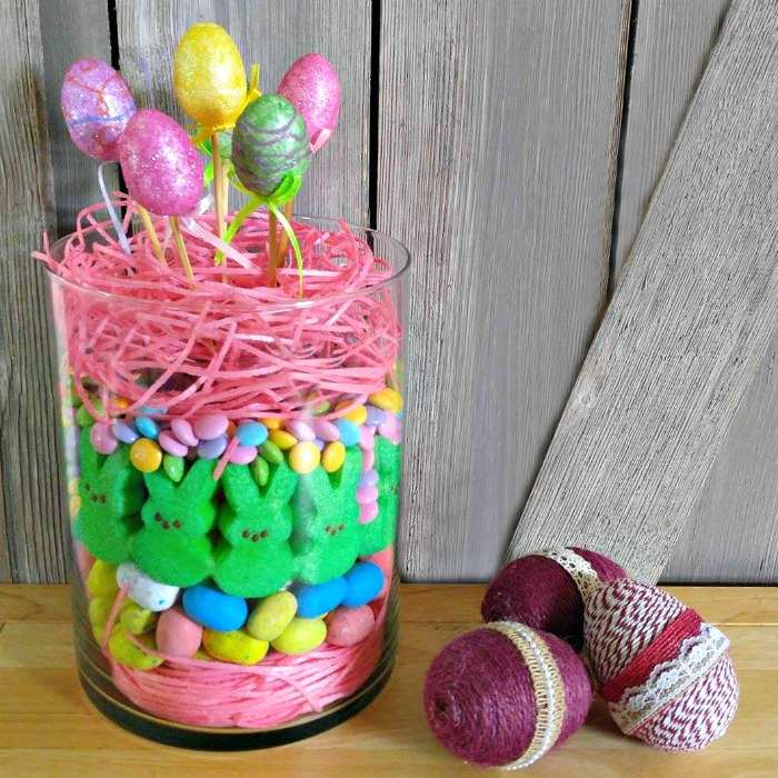 A hurricane vase filled with peeps marshmallow bunnies, robins egg candies, candy grass, and fake Easter eggs poking out of the top, it is on a tabletop next to fabric covered Easter eggs.