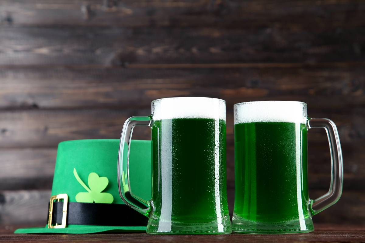 Two pints of green beer next to a green hat with black and gold buckle, and a shamrock tucked in to it against a wooden background to celebrate the symbols of St. Patrick's Day.