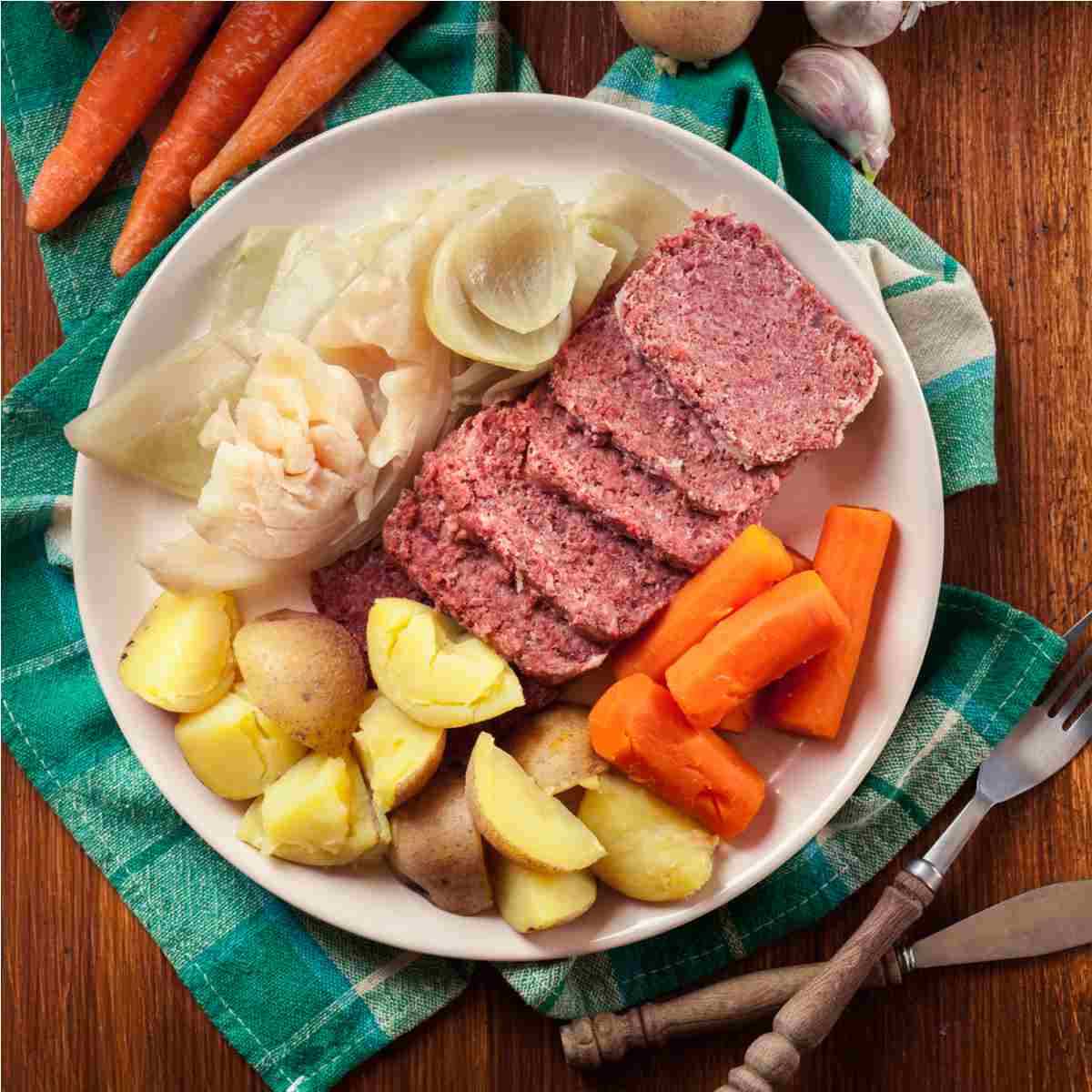 A plate of Irish corned beef and cabbage with potatoes and carrots on top of a green napkin next to a fork and knife.
