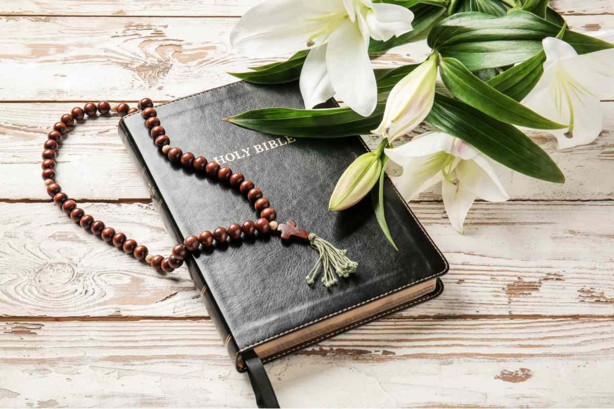 A holy bible, with a rosary and white Easter lilies arranged around it, to indicate the religious meaning of lilies in the bible.