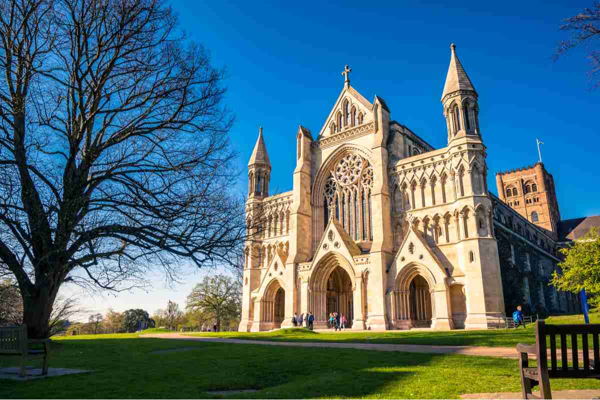 The origin location of the Alban bun, called St Albans Cathedral, against a bright blue sky, with green grass, the edge of a bench and tree without branches in the foreground of the photo.
