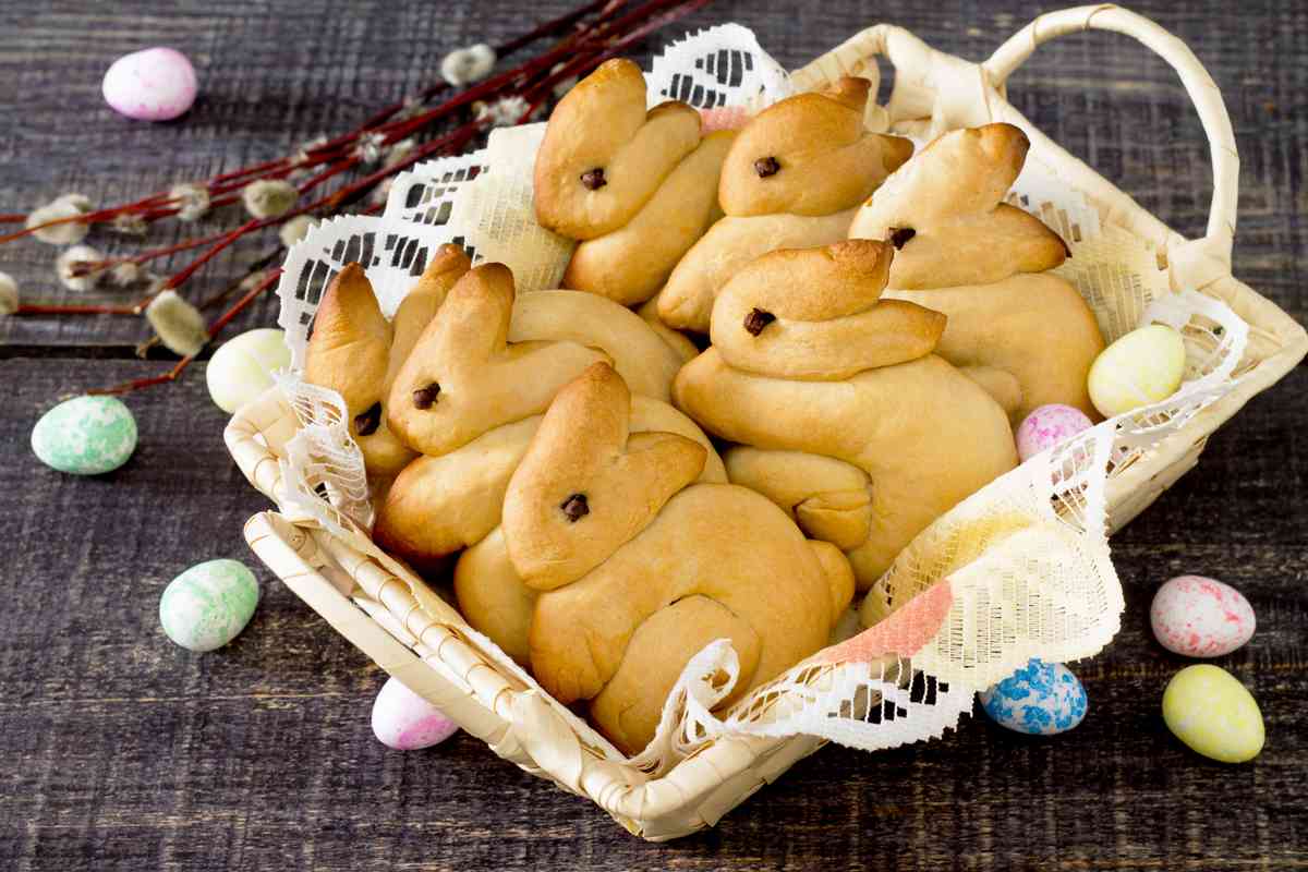 A basket of fresh baked Easter bread in the shape of bunny rabbits in a white serving tray on a doily, with robins egg candies arranged around the tray.