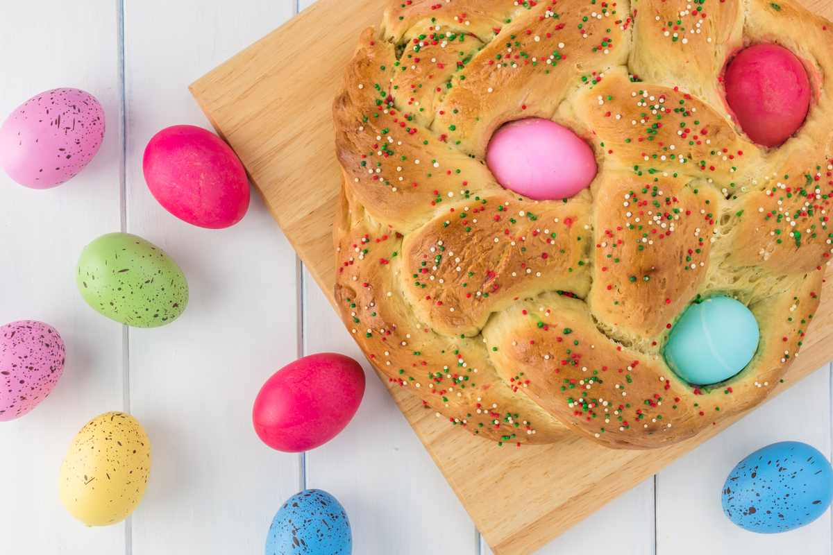 A pane di pasqua (Italian Easter bread) with dyed Eggs in it on a wooden cutting board, on top of a white wooden background with other pastel Easter eggs arranged around it.