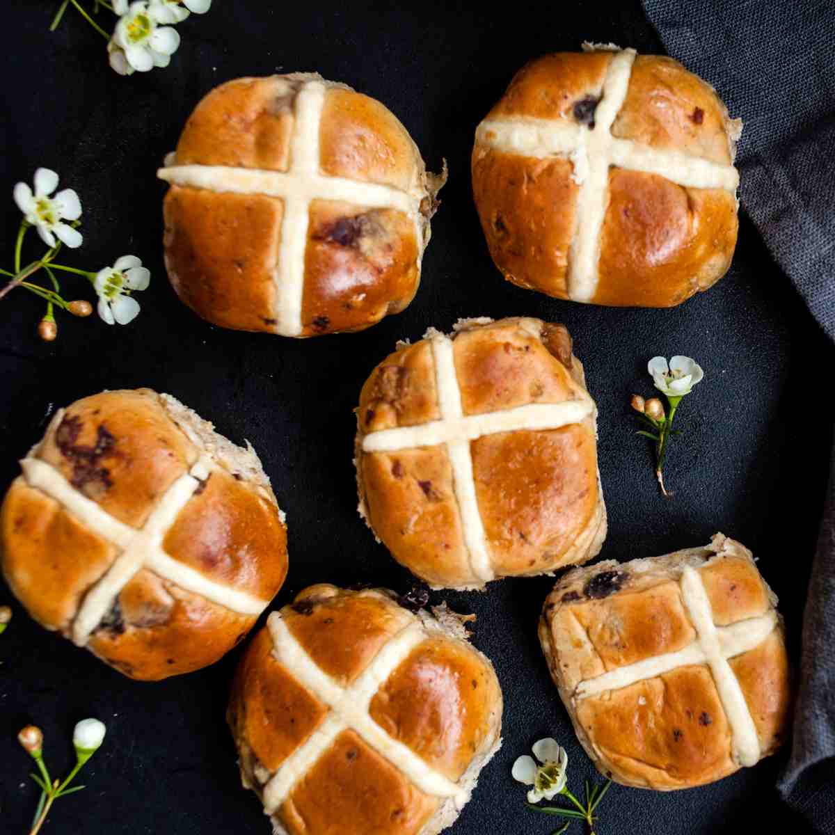 Six hot cross buns on a black napkin with tiny white flowers surrounding them, the cross prominently displayed on each of the Easter pastries symbolizing the religious hot cross buns history.