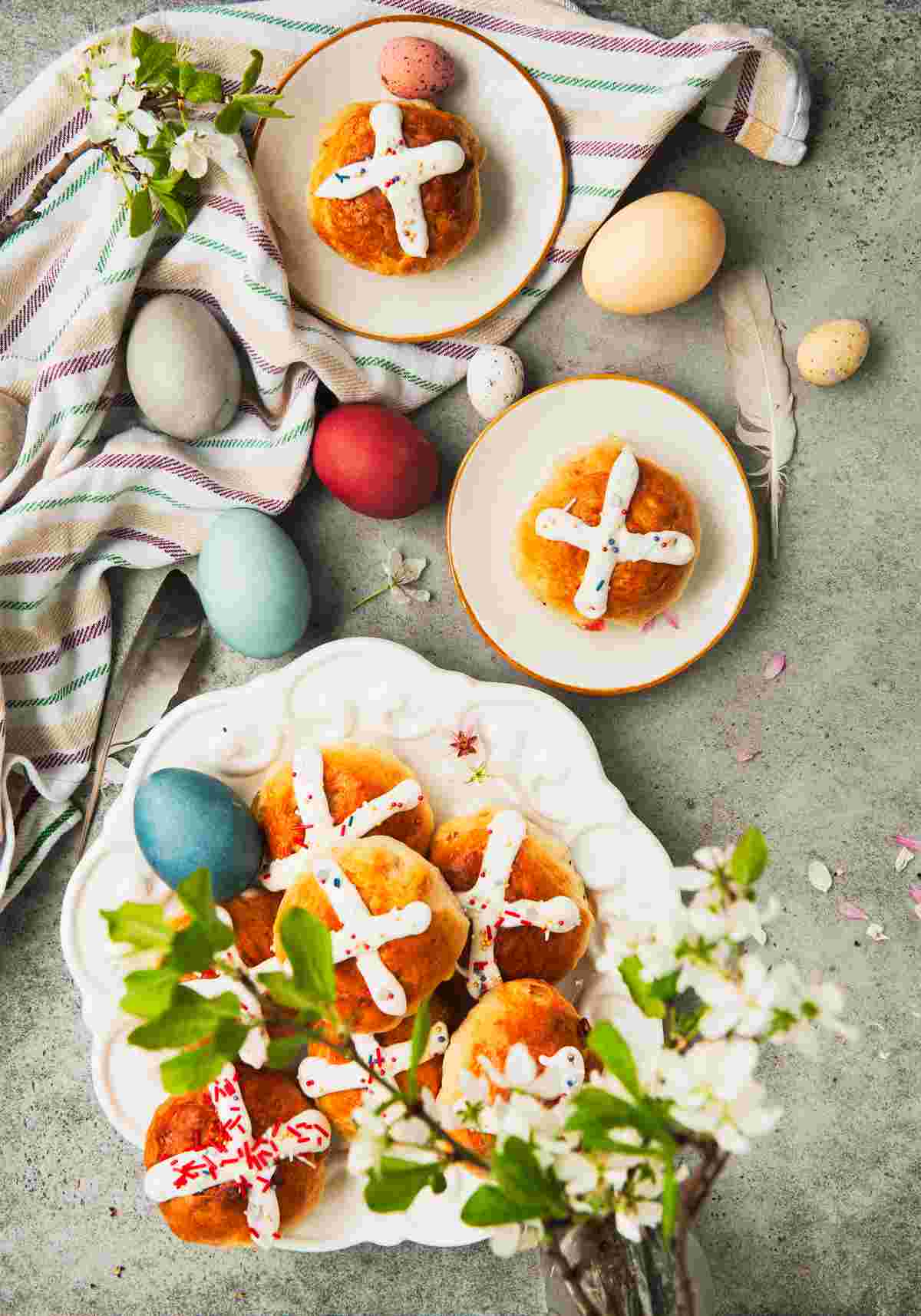Traditional hot cross buns at Easter on three separate plates, one plate containing multiple hot crossed buns, and the other two plates containing one of the Easter breads each, with Easter eggs, kitchen towels, and Easter flowers surrounding the Easter pastries. 