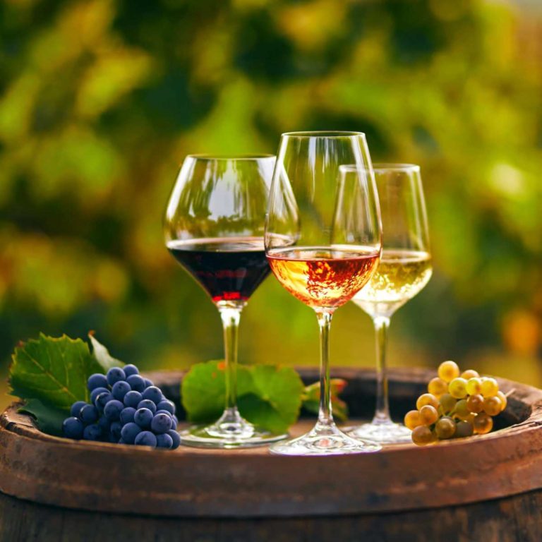 A glass of red wine, white wine and rose wine on a barrel with grapes next to them at a winery, and the wine in each glass is the ideal wine temperature.
