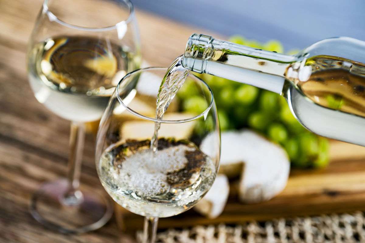 A bottle of white wine at the best white wine serving temperature being poured into two glasses on a table next to a wheel of cheese and green grapes.