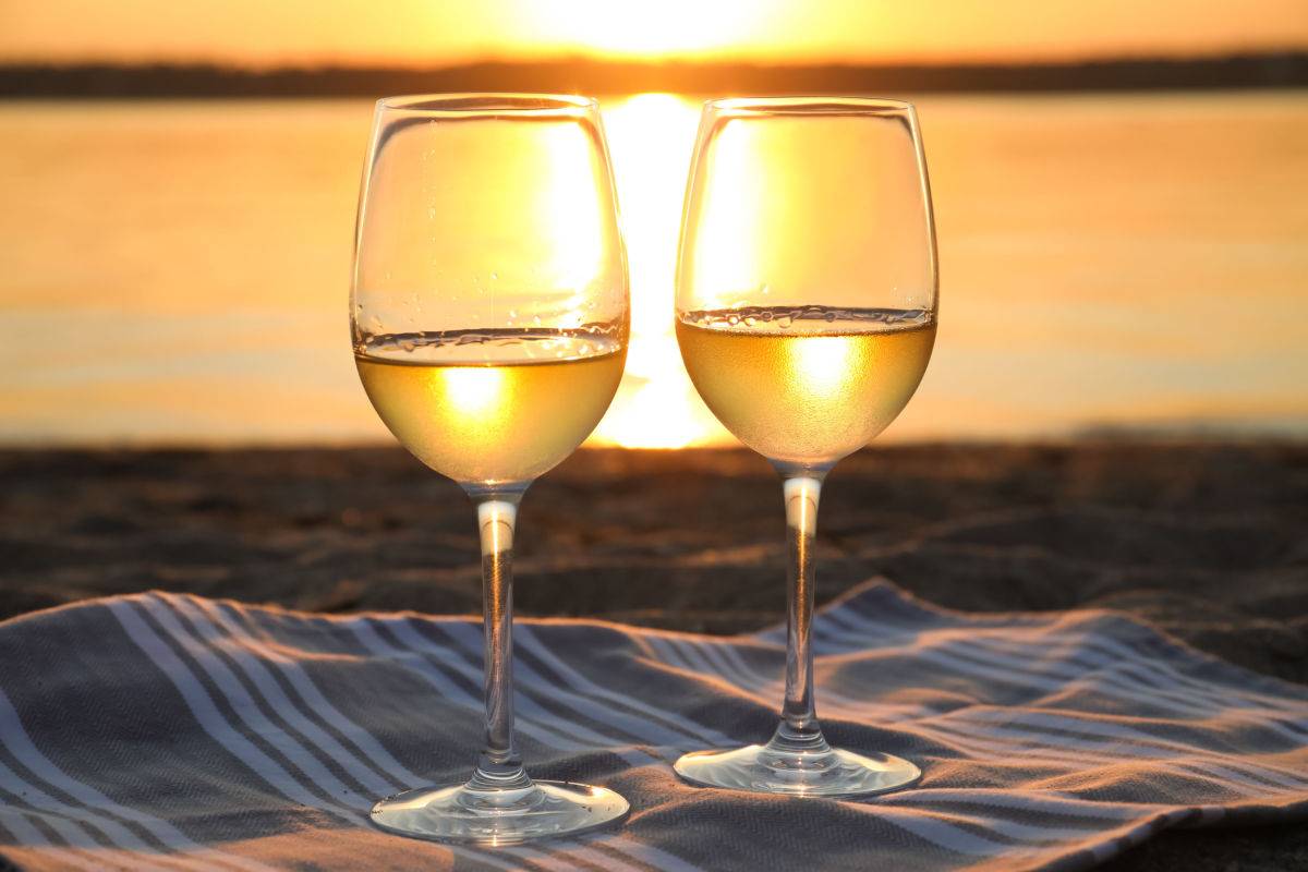 Two white wine glasses on a blanket at a beach during sunset on National White Wine Day.