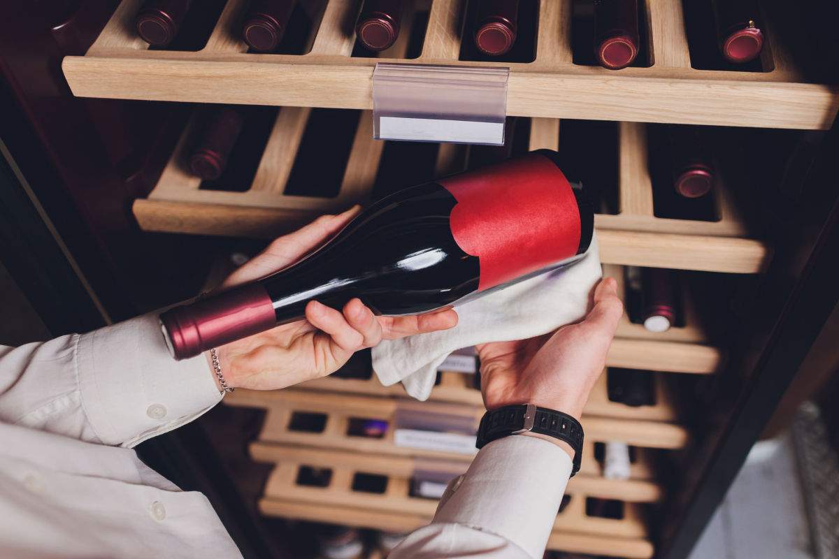 A bottle of red wine at the ideal temperature for storing red wine being pulled out of a wooden wine storage rack by someone wearing a white long sleeved button down shirt.