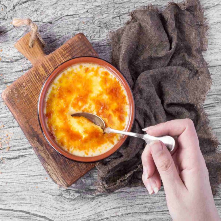 A woman's hand holding a spoon breaking the crust of a creme brulee on a cutting board with a brown towel beside it.