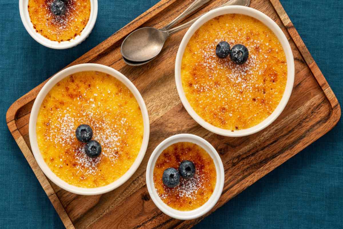 Two large creme brulee and one small one on a cutting board with two spoons, each is topped with blueberries.