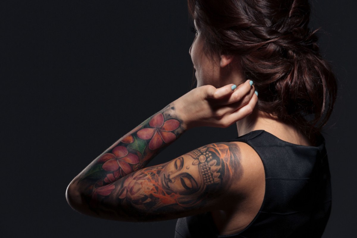 A brunette woman touching her hair with her left hand, showing off a full sleeve of tattoos on her left arm consisting of roses and a Buddha design.