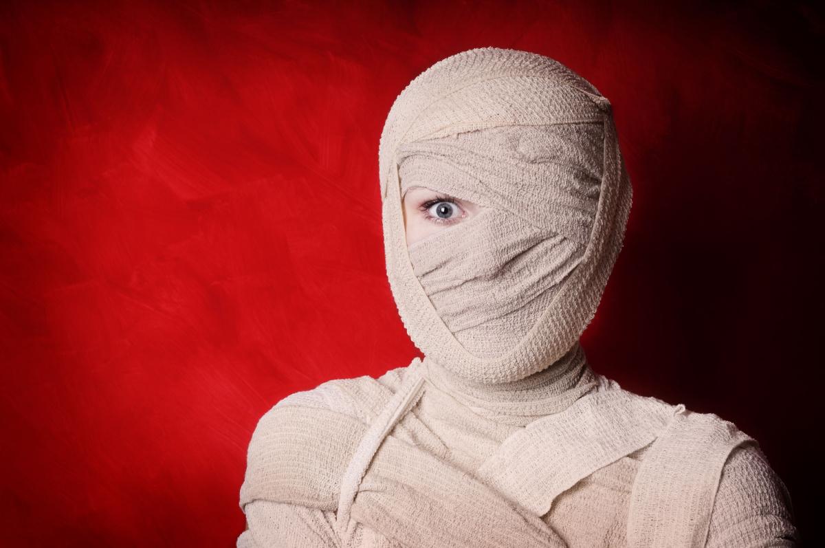 A diy mummy costume made of bandages, wrapped entirely around the person, with only her left eye visible, and she is standing in front of a red background.
