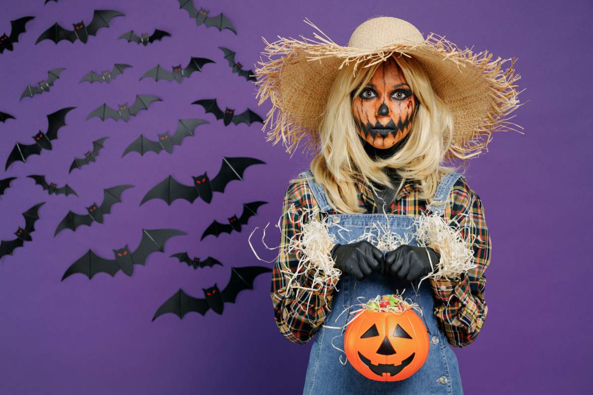 A diy scarecrow costume on a woman, consisting of denim overalls, and a plaid shirt, with straw coming out of her cuffs, and she is wearing a straw hat, with her face painted like a pumpkin, and she is holding a jack o lantern in front of a purple wall with bats.