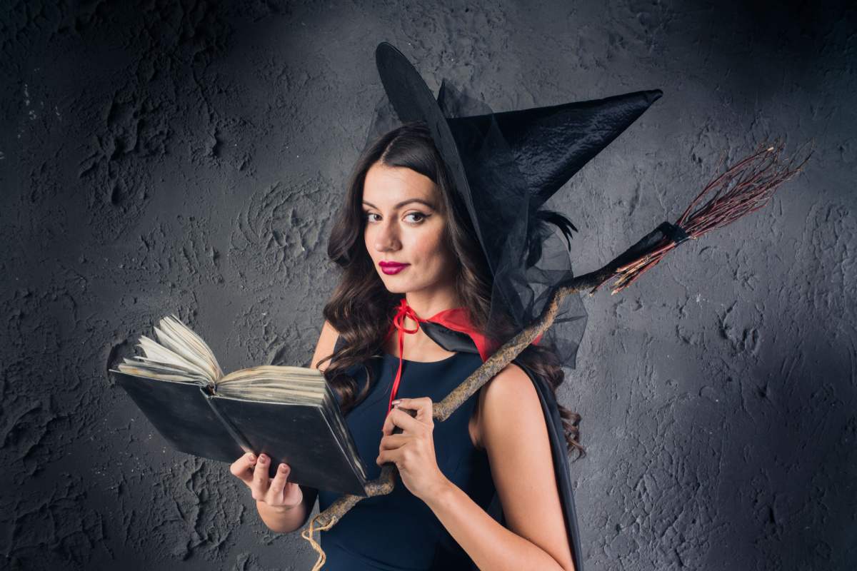 A woman wearing a DIY witch costume for Halloween consisting of a black dress, a witches hat, a black cape with a red string around her shoulders, and she is holding a broom made of sticks, and a black spell book.