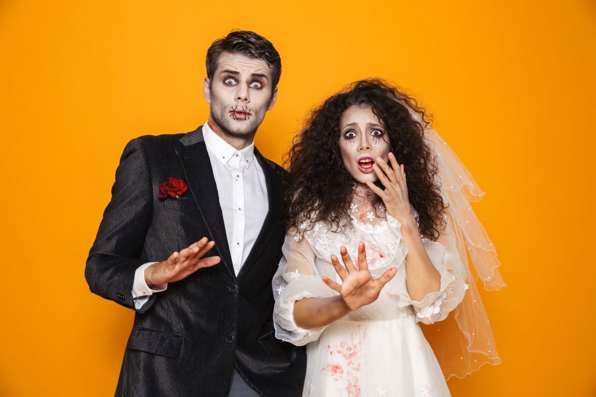 A man and woman wearing a diy zombie costume for couples, the man is in a suit with a red carnation on it, and the woman is wearing a wedding dress and veil, both have face paint to make them look like zombies.