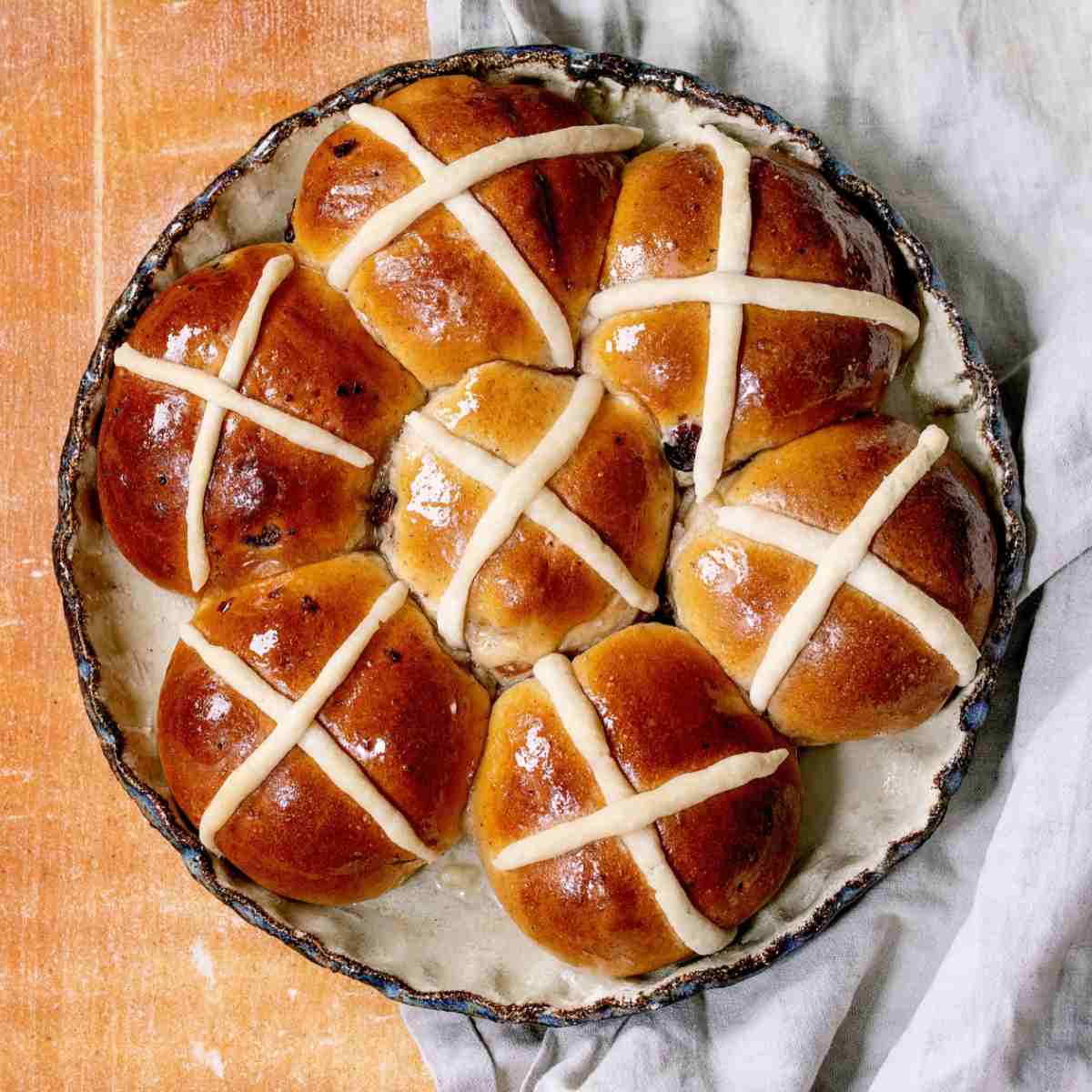 A pie tin of seven hot cross buns on a white cloth on top of a beige wooden backdrop.