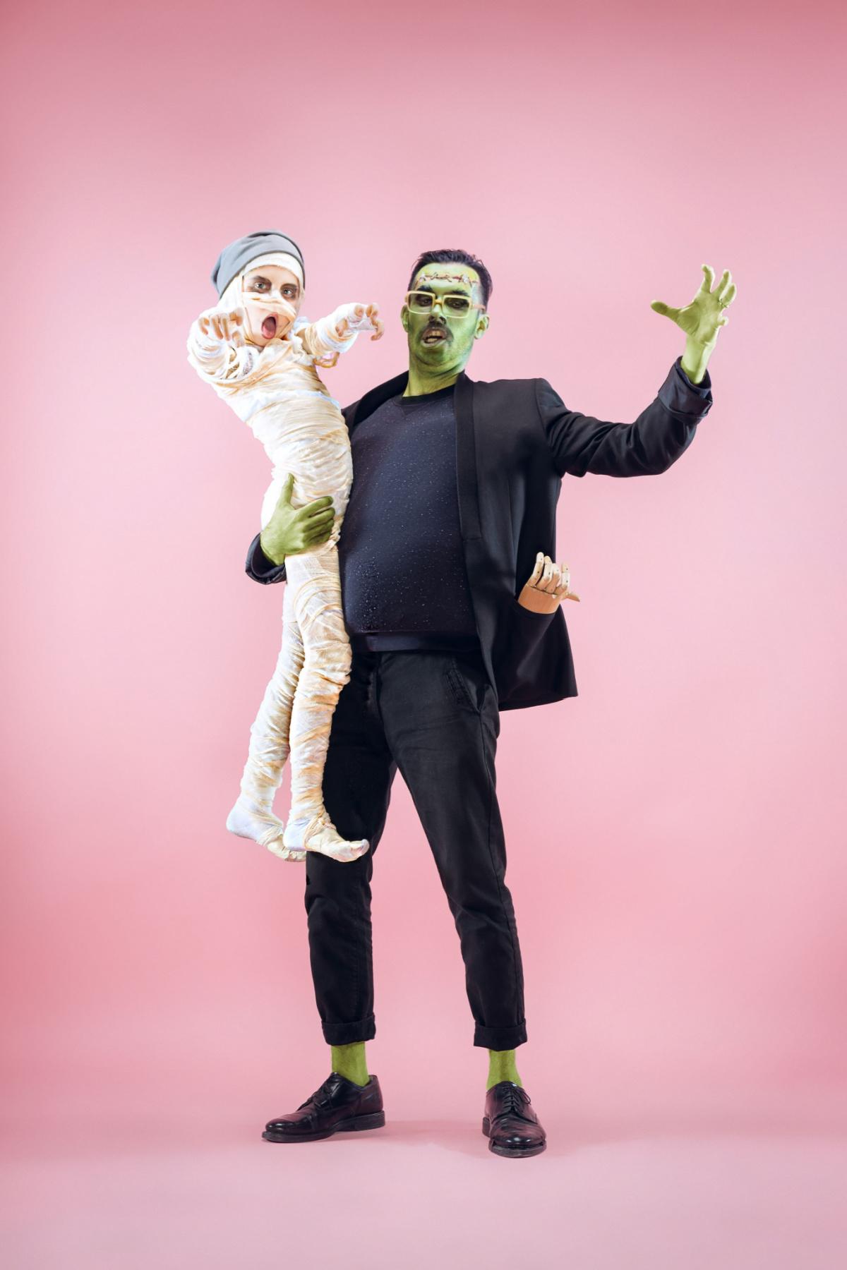 A man wearing a DIY Frankenstein costume holding a child wearing a diy mummy Halloween costume against a pink wall.
