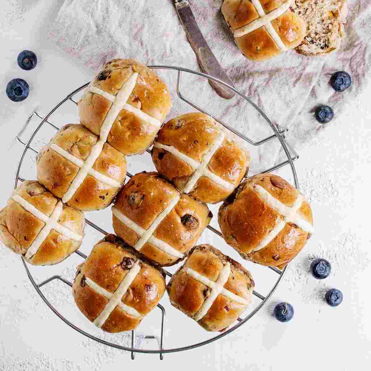 A cooling rack of eight hot cross buns on National Hot Cross Bun Day with one hot cross bun on a cloth beside it, all of them are surrounded by blueberries.