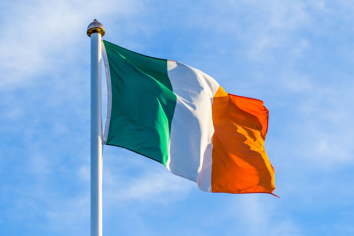 A tricolor Irish flag composed of the Irish flag colors, in order: green, white and orange vertical stripes of equal size attached to a flagstaff, blowing in the wind.