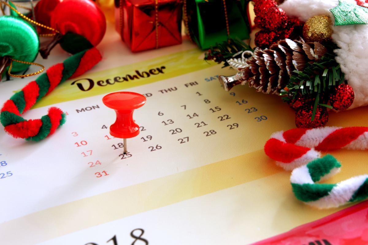A calendar of December with a red push pin on December 25 on a table with crafting supplies on top including felt candy canes, glittery pine cones, and tiny presents.