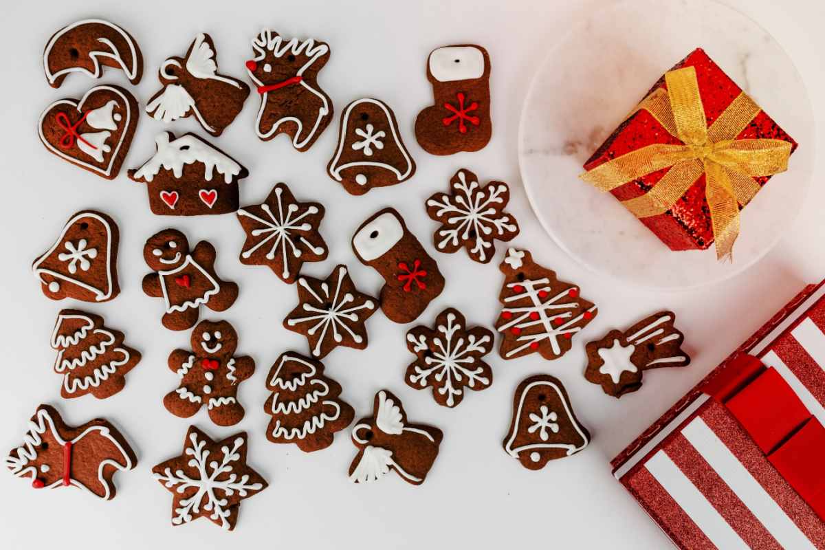 Gingerbread cookies in various festive shapes like snowflakes, snowmen, stars, reindeer, angels, bells, and stockings on a white table next to a small red present with a golden bow on it.