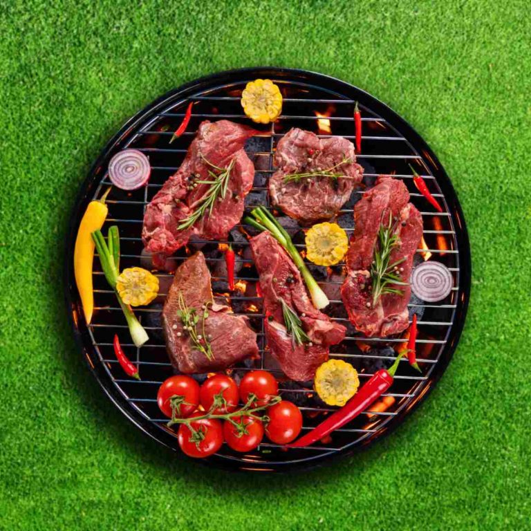 A charcoal grill on a green lawn with raw steaks and veggies cooking on the grill grate for National Grilling Day.