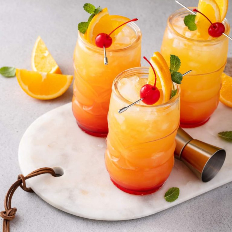 Three mai tai rum cocktails on a marble cutting board, each one is garnished with an orange slice and maraschino cherry on top. with o