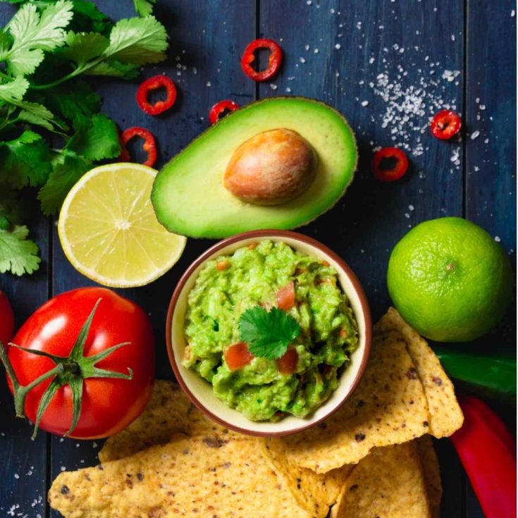 A bowl of guacamole with guacamole ingredients around it including avocados, lime, tomato, cilantro, serrano pepper, lemon and salt.