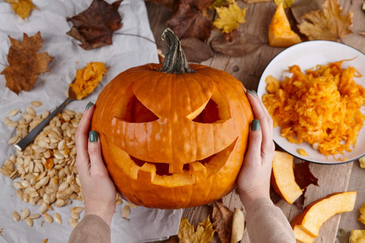A person's hands holding a carved pumpkin with a jack o lantern face surrounded by leaves, pumpkin seeds and pumpkin pulp.